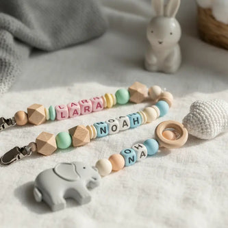 Zwei handgefertigte, personalisierte Schnullerketten aus Holzperlen mit dem Namen Noah und einem grauen Elefanten-Anhänger auf hellem Stoffhintergrund.