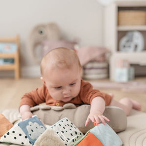 Baby liegt auf dem Bauch und spielt mit einem ausklappbaren Stoffwürfel in bunten Farben – fördert Motorik und Sinne, erhältlich bei BabyLions.