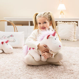 Mädchen sitzt fröhlich auf einem weichen Teppich und kuschelt mit einem weißen Einhorn-Plüschtier aus der Babylions Kollektion.