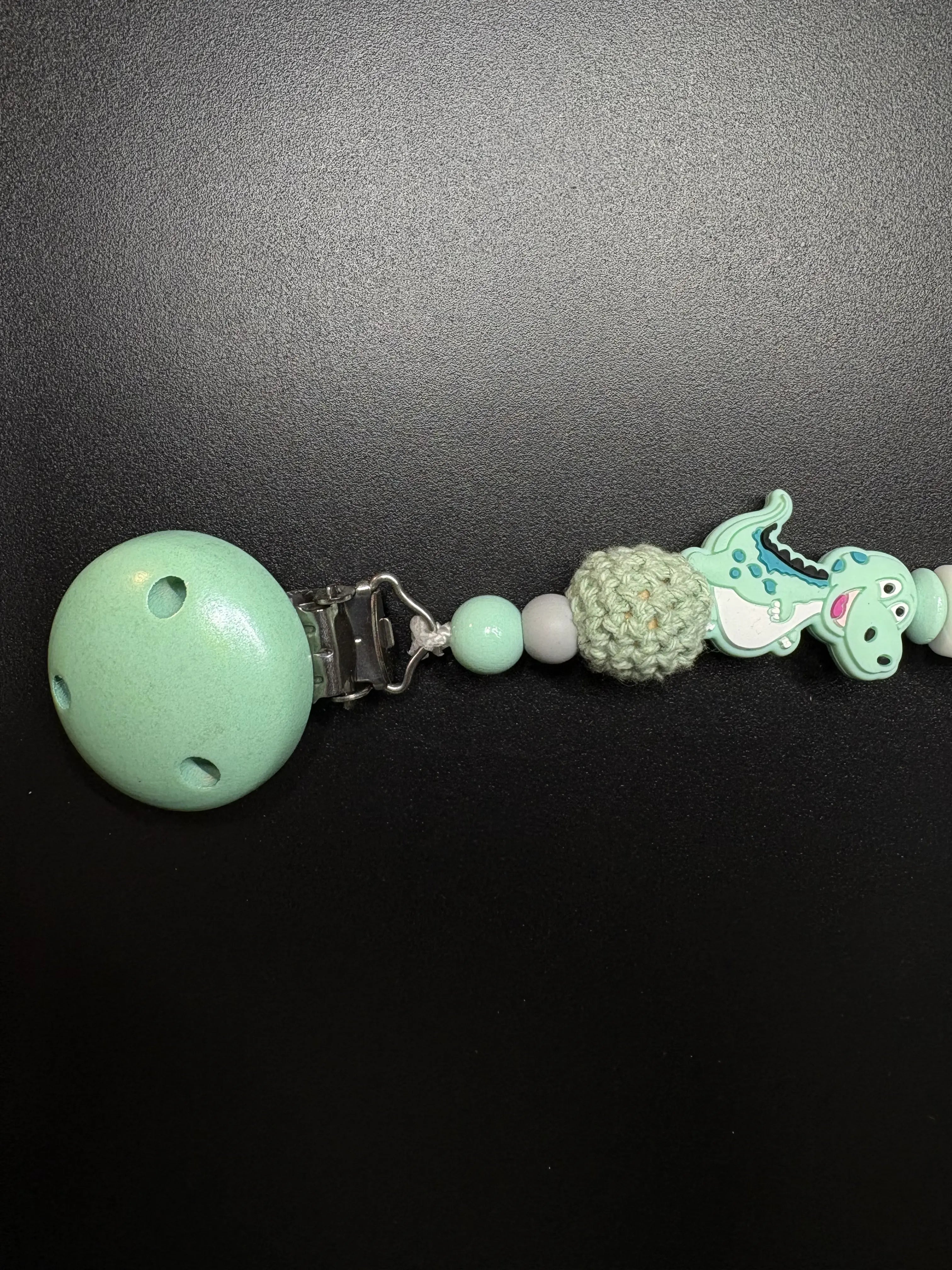 Seitenansicht der personalisierten Schnullerkette Dino mit Namen von BabyLions® mit Dino-Motiv, gehäkelter Perle, Holzclip und hochwertigen Silikon- sowie Holzperlen in Mint