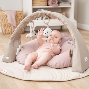 Baby liegt auf einem rosafarbenen Kissen unter einem beigen Spielbogen mit weichen Hängespielzeugen in Tierform – fördert Motorik und Sinnesentwicklung, erhältlich bei BabyLions.