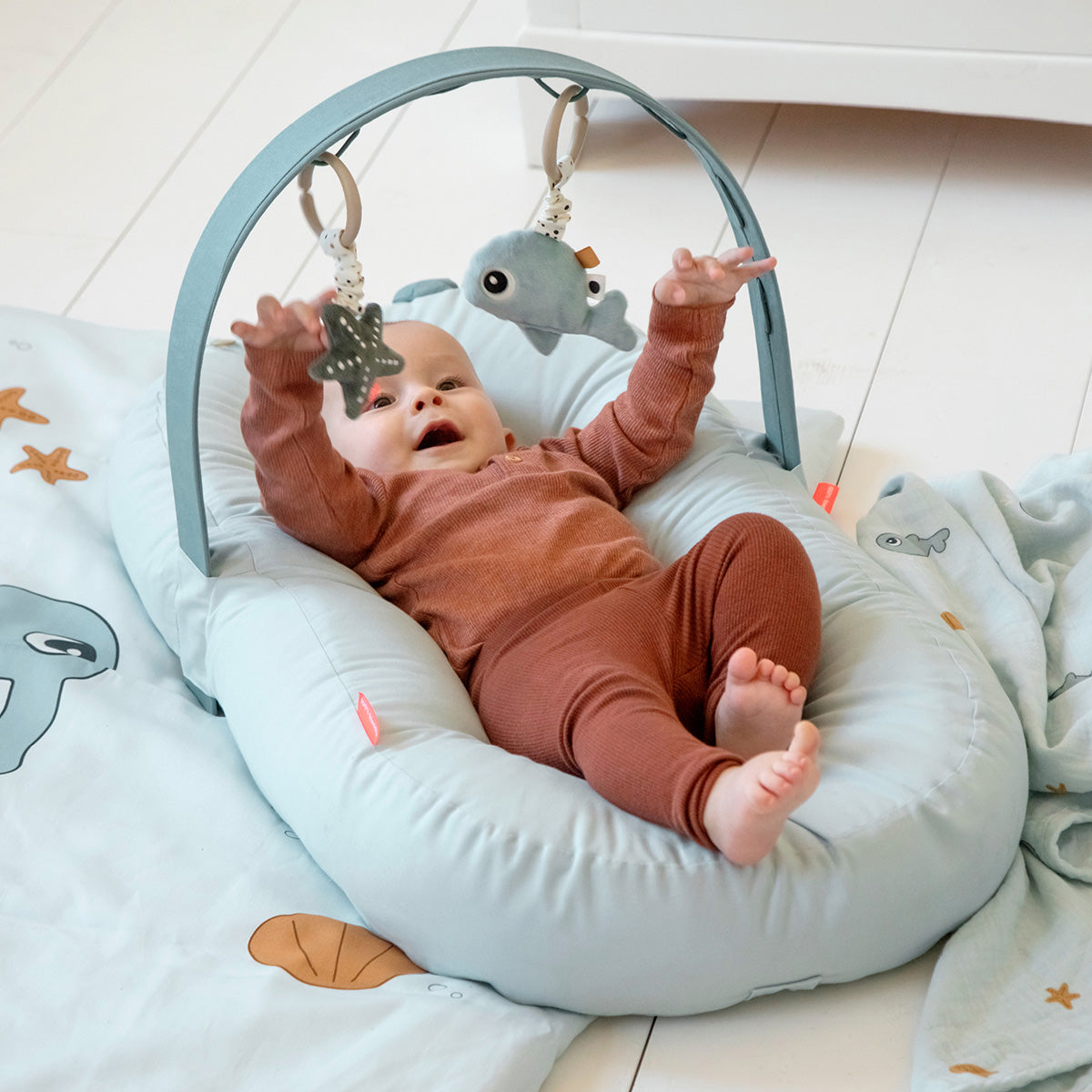 Baby liegt im Babynest mit Spielbogen und Greifspielzeug – Aktivitätsspielzeug für Babys mit Rassel und Spiegel in Blau
