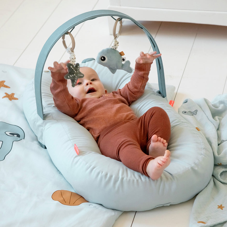 Baby liegt im Babynest mit Spielbogen und Greifspielzeug – Aktivitätsspielzeug für Babys mit Rassel und Spiegel in Blau