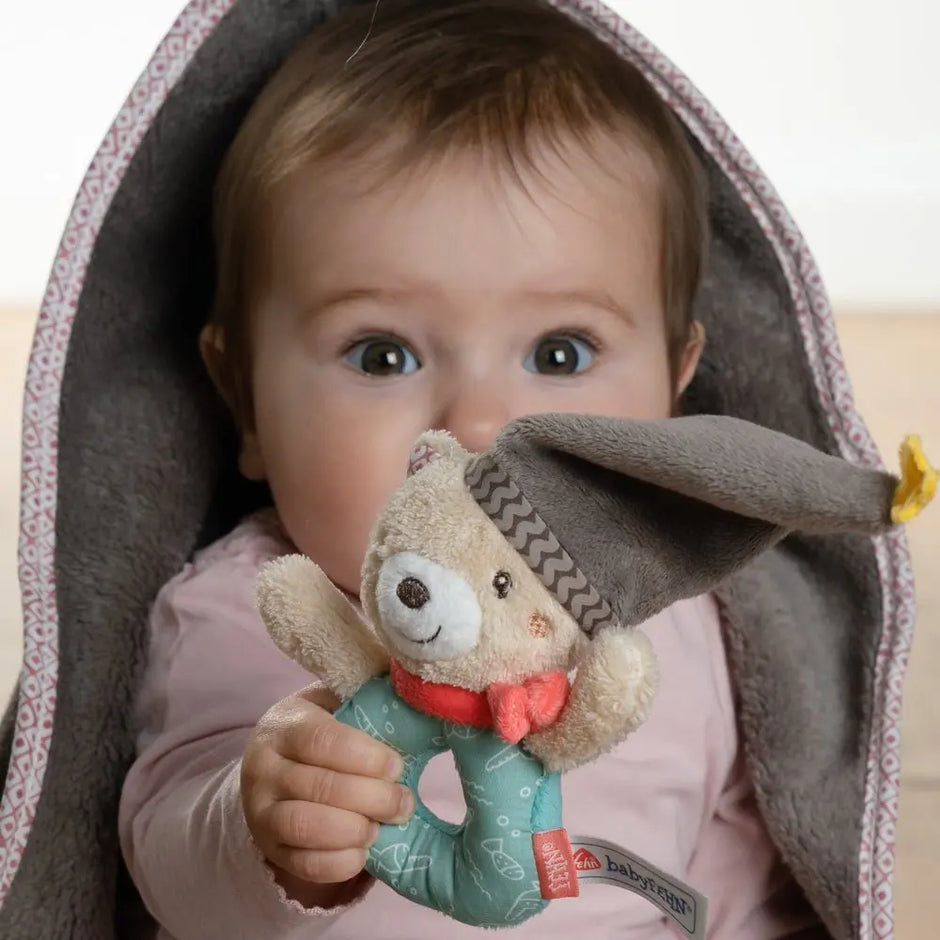 Baby Kuscheldecke Bär grau mit Greifring – Baby hält Spielzeug und schaut unter der Decke hervor