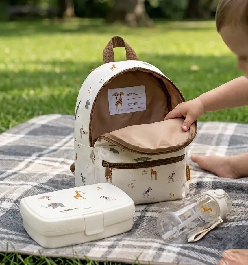 Kind öffnet einen Kinderrucksack im Safari-Design mit passender Brotdose und Trinkflasche auf einer Picknickdecke im Freien.