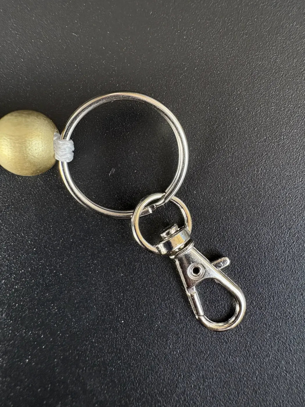 Nahaufnahme des silberfarbenen Schlüsselrings mit Karabinerhaken eines personalisierten BabyLions® Schlüsselanhängers Reh, befestigt an einer goldfarbenen Holzperle mit weißer Kordel auf schwarzem Hintergrund.