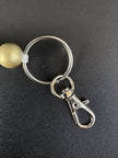 Nahaufnahme des silberfarbenen Schlüsselrings mit Karabinerhaken eines personalisierten BabyLions® Schlüsselanhängers Reh, befestigt an einer goldfarbenen Holzperle mit weißer Kordel auf schwarzem Hintergrund.