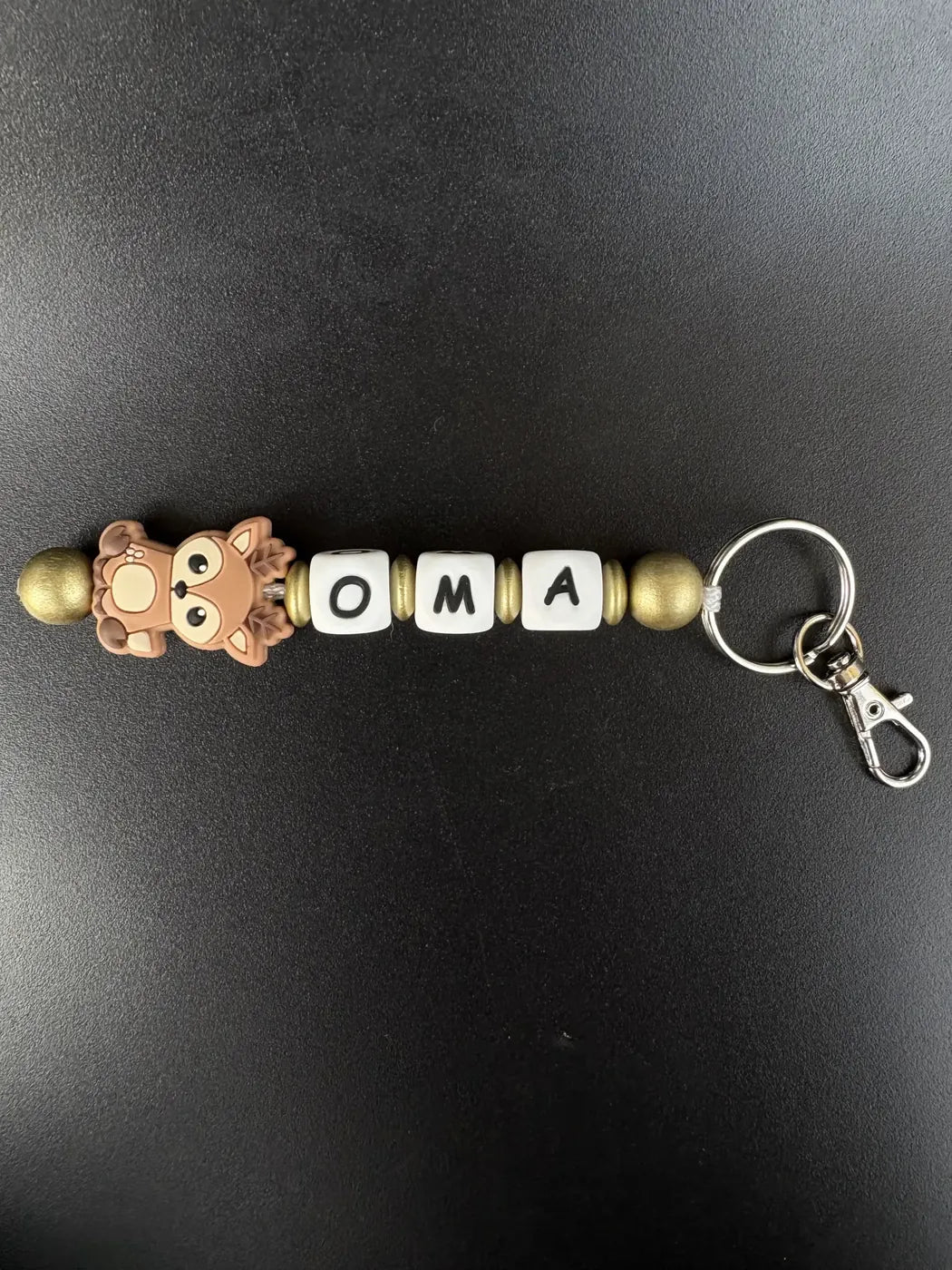 Personalisierter Schlüsselanhänger Reh von BabyLions® mit dem Schriftzug OMA, niedlichem Rehmotiv, goldfarbenen Perlen und Metallring mit Karabiner auf schwarzem Hintergrund.