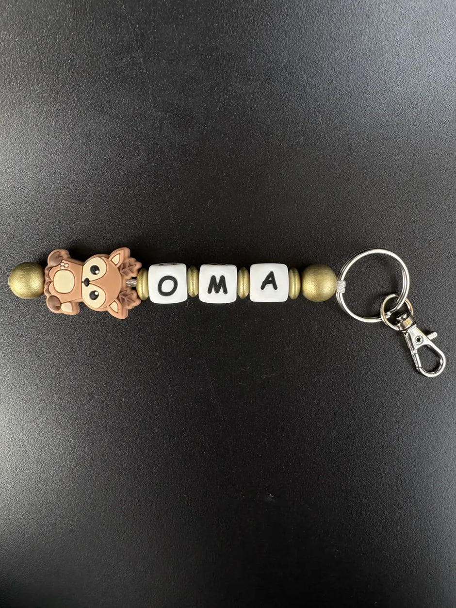 Personalisierter Schlüsselanhänger Reh von BabyLions® mit dem Schriftzug OMA, niedlichem Rehmotiv, goldfarbenen Perlen und Metallring mit Karabiner auf schwarzem Hintergrund.
