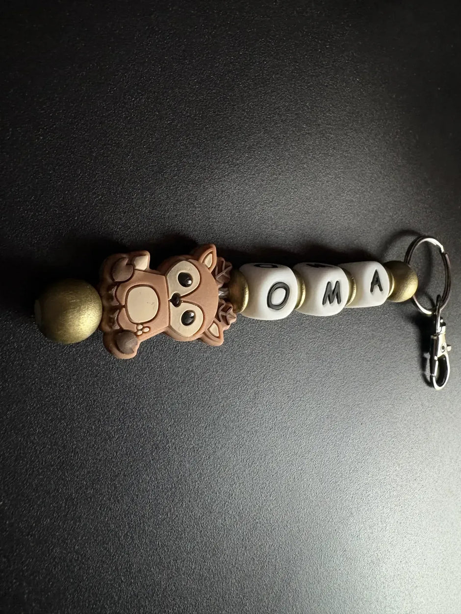 Handgefertigter personalisierter BabyLions® Schlüsselanhänger mit niedlichem Reh-Motiv, weißen Buchstabenperlen mit dem Namen OMA, goldfarbenen Holzperlen und silbernem Schlüsselring mit Karabiner auf schwarzem Hintergrund.