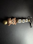 Handgefertigter personalisierter BabyLions® Schlüsselanhänger mit niedlichem Reh-Motiv, weißen Buchstabenperlen mit dem Namen OMA, goldfarbenen Holzperlen und silbernem Schlüsselring mit Karabiner auf schwarzem Hintergrund.