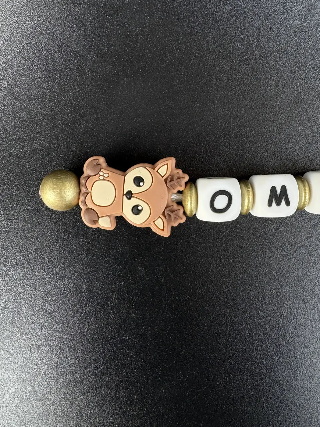 Detailaufnahme eines personalisierten BabyLions® Schlüsselanhängers mit niedlichem braunem Rehmotiv, weißen Buchstabenperlen mit dem Schriftzug OMA und goldfarbenen Perlen auf schwarzem Hintergrund.