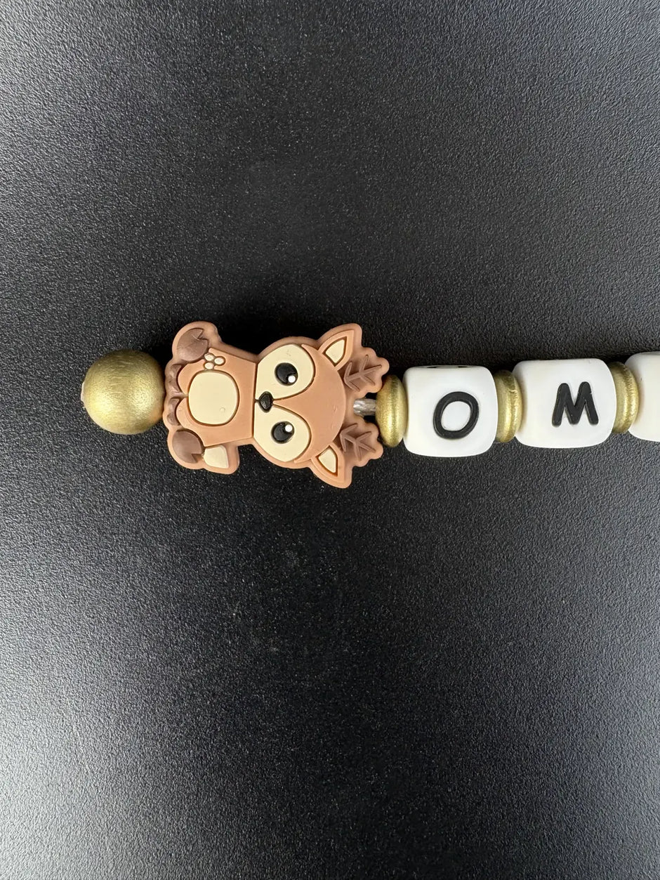 Detailaufnahme eines personalisierten BabyLions® Schlüsselanhängers mit niedlichem braunem Rehmotiv, weißen Buchstabenperlen mit dem Schriftzug OMA und goldfarbenen Perlen auf schwarzem Hintergrund.