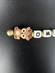 Detailaufnahme eines personalisierten BabyLions® Schlüsselanhängers mit niedlichem braunem Rehmotiv, weißen Buchstabenperlen mit dem Schriftzug OMA und goldfarbenen Perlen auf schwarzem Hintergrund.