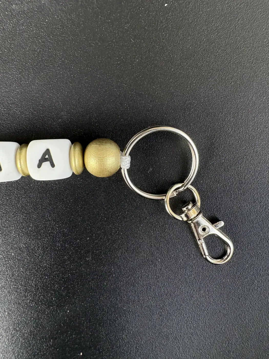 Detailaufnahme des personalisierten BabyLions® Schlüsselanhängers Reh mit silberfarbenem Schlüsselring, Karabinerhaken, goldfarbenen Perlen und weißer Buchstabenperle auf schwarzem Hintergrund.