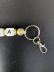 Detailaufnahme des personalisierten BabyLions® Schlüsselanhängers Reh mit silberfarbenem Schlüsselring, Karabinerhaken, goldfarbenen Perlen und weißer Buchstabenperle auf schwarzem Hintergrund.