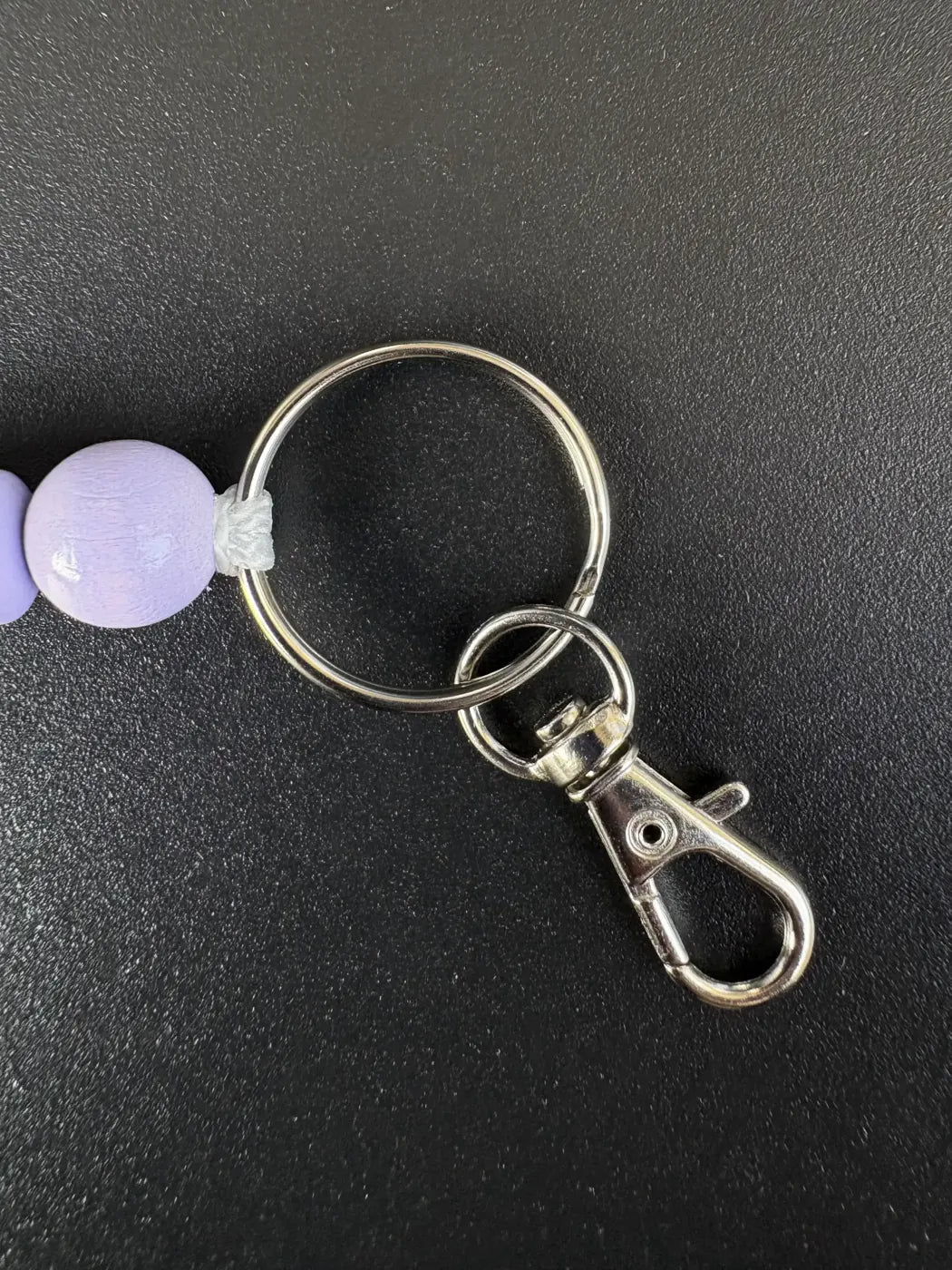 Nahaufnahme des silberfarbenen Schlüsselrings mit Karabiner und fliederfarbener Holzperle eines handgefertigten BabyLions® Schlüsselanhängers Schmetterling auf schwarzem Hintergrund.