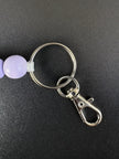 Nahaufnahme des silberfarbenen Schlüsselrings mit Karabiner und fliederfarbener Holzperle eines handgefertigten BabyLions® Schlüsselanhängers Schmetterling auf schwarzem Hintergrund.