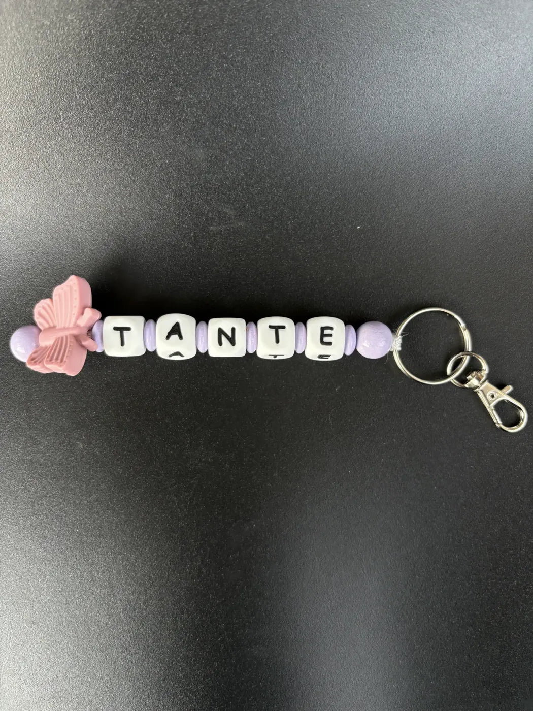 Handgefertigter personalisierter BabyLions® Schlüsselanhänger mit rosa Schmetterling, weißen Buchstabenperlen mit der Aufschrift TANTE, fliederfarbenen Perlen und silbernem Schlüsselring mit Karabiner auf schwarzem Hintergrund.