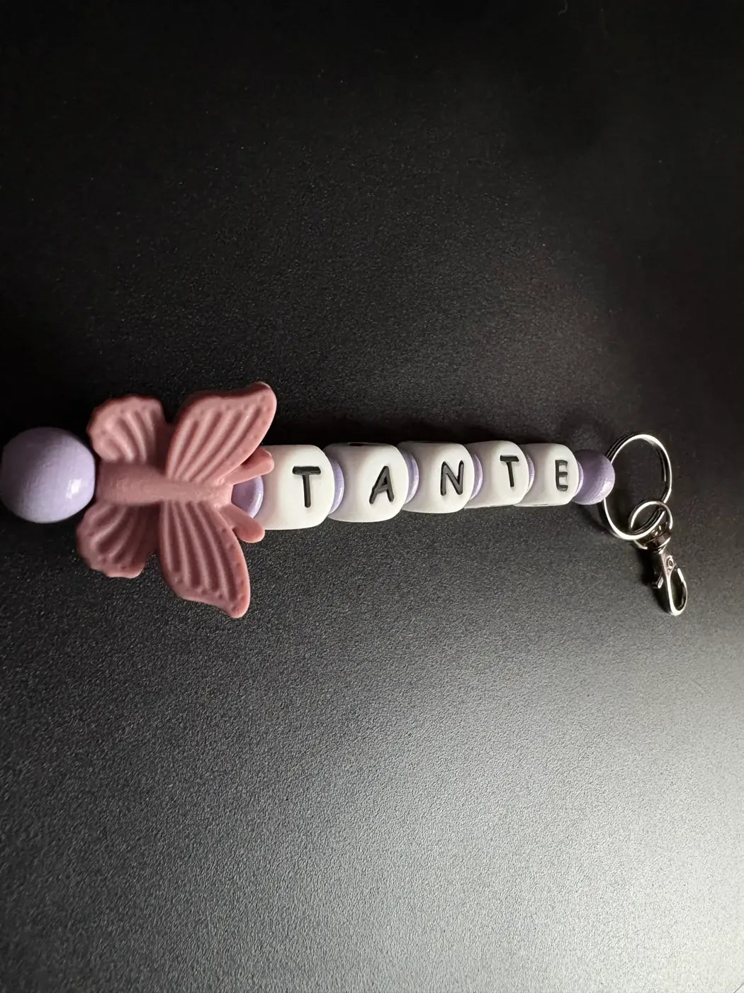 Handgefertigter BabyLions® Schlüsselanhänger mit rosa Schmetterling, fliederfarbenen Perlen und weißen Buchstabenwürfeln mit der Aufschrift TANTE. Vollständige Ansicht mit silbernem Schlüsselring und Karabiner auf schwarzem Hintergrund.