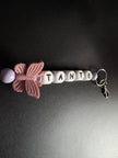 Handgefertigter BabyLions® Schlüsselanhänger mit rosa Schmetterling, fliederfarbenen Perlen und weißen Buchstabenwürfeln mit der Aufschrift TANTE. Vollständige Ansicht mit silbernem Schlüsselring und Karabiner auf schwarzem Hintergrund.