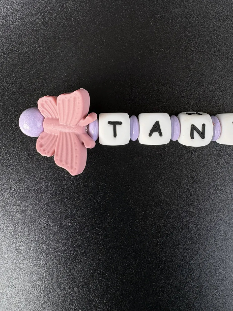 Nahaufnahme eines handgefertigten BabyLions® Schlüsselanhängers mit rosa Schmetterling, fliederfarbenen Perlen und weißen Buchstabenwürfeln mit schwarzer Schrift. Detailansicht des personalisierten Schlüsselanhängers auf schwarzem Hintergrund.