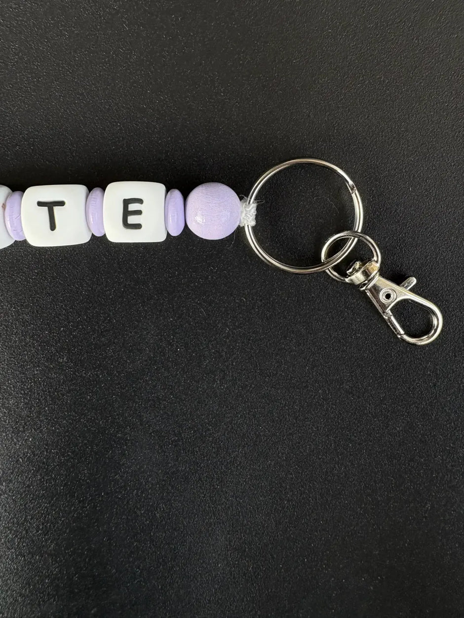 Detailaufnahme eines personalisierten BabyLions® Schlüsselanhängers Schmetterling mit fliederfarbenen Perlen, weißen Buchstabenwürfeln sowie silbernem Schlüsselring und Karabiner auf schwarzem Hintergrund.