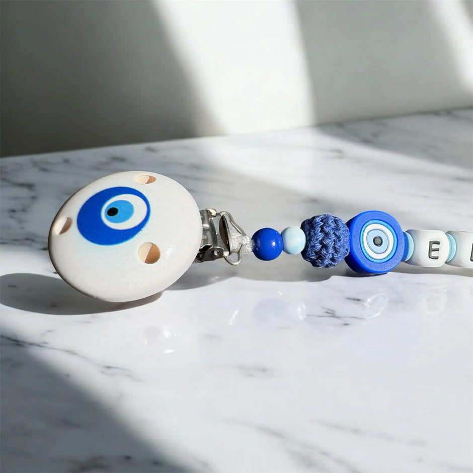 Detailaufnahme einer Schnullerkette mit blauem Nazar Auge, Holzclip und Silikonperlen in Blau und Weiß auf hellem Marmortisch