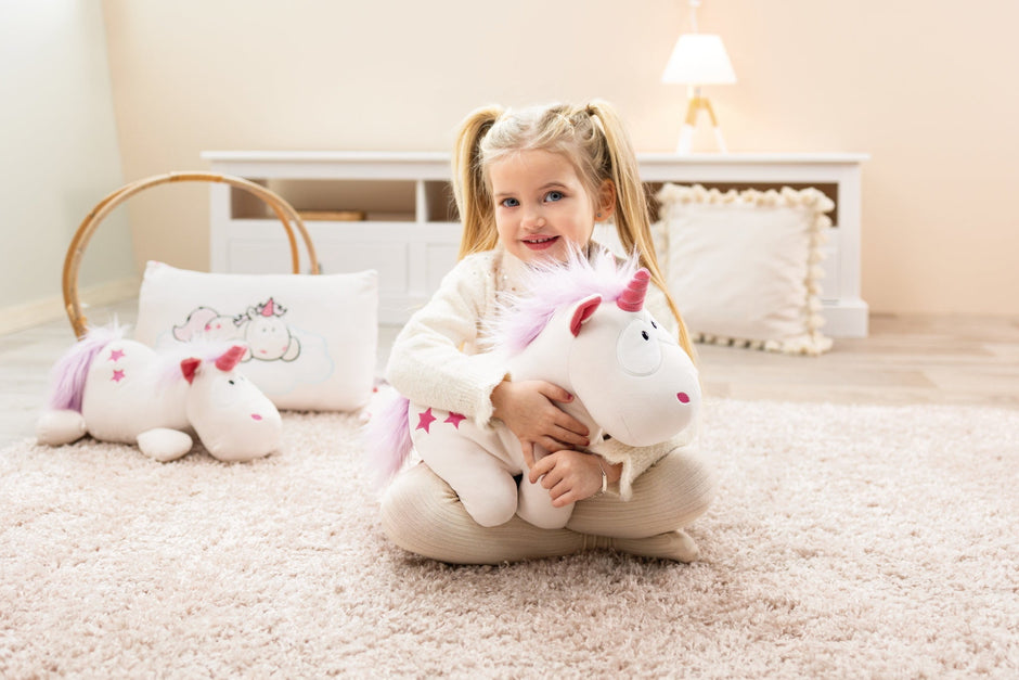 Mädchen mit blonden Zöpfen sitzt auf einem weichen Teppich und umarmt ein weiß-pinkes Kuscheltier-Einhorn „Theodor“ mit rosa Mähne und Sternen – erhältlich bei BabyLions.