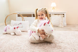 Mädchen mit blonden Zöpfen sitzt auf einem weichen Teppich und umarmt ein weiß-pinkes Kuscheltier-Einhorn „Theodor“ mit rosa Mähne und Sternen – erhältlich bei BabyLions.
