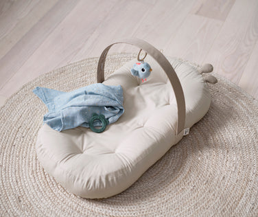 Beige Cozy Lounger mit Holz-Spielbogen, maritimen Hängespielzeugen und weicher Babydecke – gemütlicher Spiel- und Ruheplatz für Neugeborene.