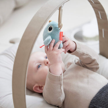 Baby liegt auf einem weichen Cozy Lounger und spielt mit einem hängenden Stofftier in Meeresform unter einem Holz-Spielbogen.