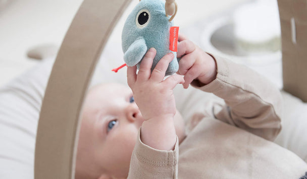 Baby liegt auf einem weichen Cozy Lounger und spielt mit einem hängenden Stofftier in Meeresform unter einem Holz-Spielbogen.